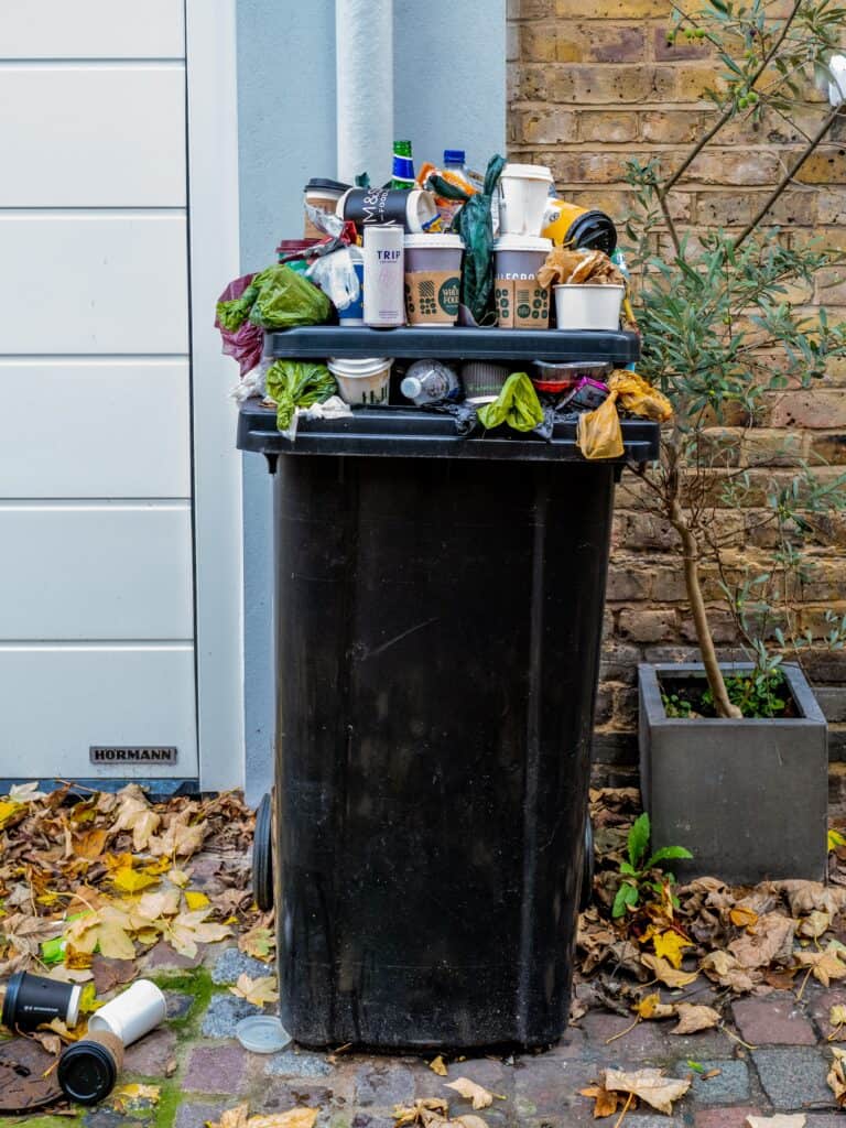 overfull black garbage can sitting in front of white garage door and brick house